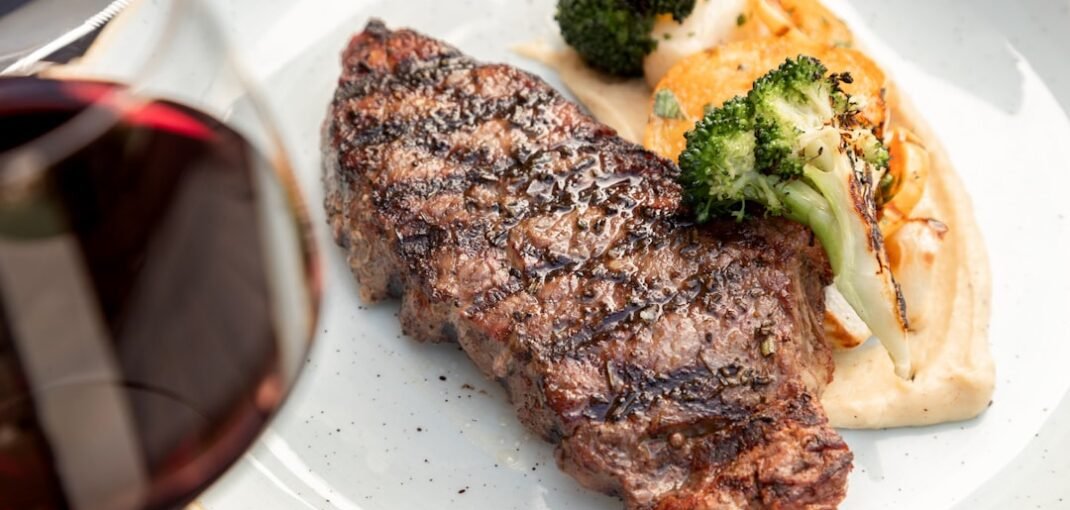 A glass of red wine and a whisky beside a perfectly seared steak on a wooden board, whisky wine gin steak pairings